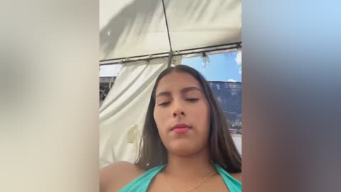 Video of a young woman with medium brown skin and long, straight dark brown hair, wearing a green halter top, standing in front of a white tent with a mountainous landscape visible through the tent's windows.