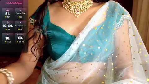 Video of a woman in a teal sari with gold embroidery and a sheer white dupatta adorned with golden dots, wearing a gold necklace. Background shows a smartphone screen displaying \"Lovemee\" app with stats.