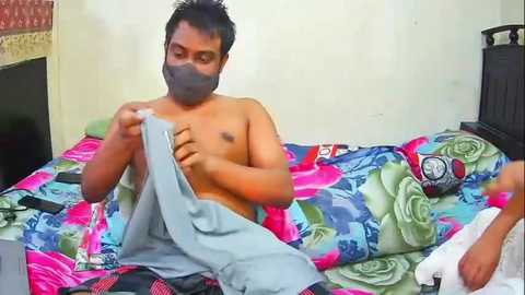 Video of a shirtless man wearing a face mask, adjusting a gray blanket on a colorful floral bedspread in a modest bedroom.