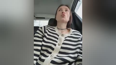 Media: Video of a woman in a car, wearing a black and white striped shirt, black choker, and shoulder harness, with a neutral expression.