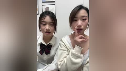 Video of two young Asian women in white blouses, one with a red bow tie and the other with a cellphone, standing in a white-walled room with framed art.