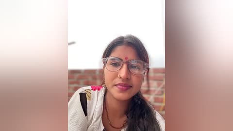 A video of a young South Asian woman with medium brown skin, wearing glasses and a traditional white top with embroidery, against a brick wall background.