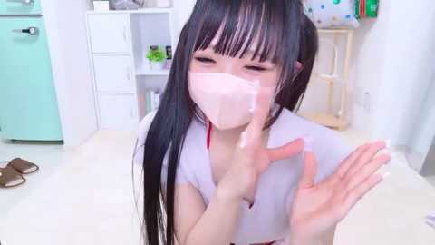 Media: Video of a young Asian woman with long black hair and fair skin, wearing a white mask, holding hands to her face, in a brightly lit room with white walls and pastel furniture.