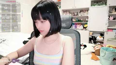 A video of a young woman with straight black hair, wearing a striped tank top, seated in a gaming chair in a cluttered room filled with stuffed animals and gaming equipment.