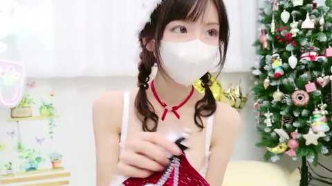 Video of a young Asian woman with braided hair, wearing a white face mask, red choker, and a white spaghetti-strap dress, holding a black microphone, standing in front of a decorated Christmas tree.