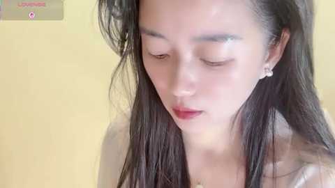 Video of an Asian woman with long, dark hair, wearing a white top, and subtle makeup, against a pale yellow background.