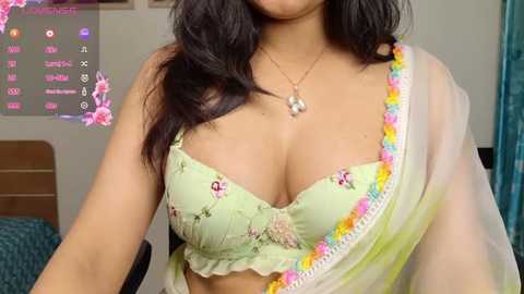 Media: Video of a light-skinned woman with long, wavy dark hair, wearing a pastel green bra with pink floral embroidery and a sheer, multicolored sari, set against a modern bedroom with a digital frame displaying social media icons.