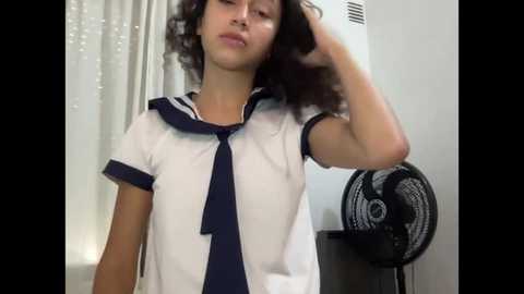 Media: A video of a young woman with curly hair, wearing a Japanese schoolgirl uniform, adjusting her hair in front of a white wall and a black fan.