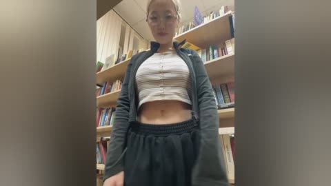 Video of a fair-skinned woman with blonde hair in a striped crop top, black hoodie, and baggy black pants, standing in a well-lit, book-filled room.