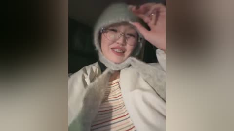 A video of a smiling Asian woman with glasses, wearing a white hat, scarf, and striped shirt, adjusting her hat in a dimly lit room.
