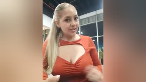 Video of a blonde woman with a fair complexion wearing a tight, low-cut orange top, smiling and gesturing, indoors with a modern background.