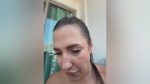 Video of a fair-skinned woman with wet hair, wearing minimal makeup, looking down thoughtfully. Background features a green shower curtain and a partially visible window.