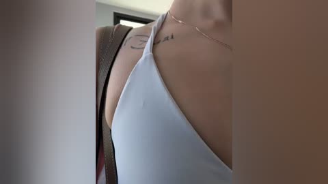 Video of a woman's bare shoulder and upper chest in a white tank top, revealing a tattoo. She wears a delicate gold necklace. Background shows a blurred, modern interior with a window.