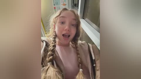 A video of a young, fair-skinned girl with long, blonde braids, wearing a light pink top and brown jacket, smiling widely while peering out of a window.