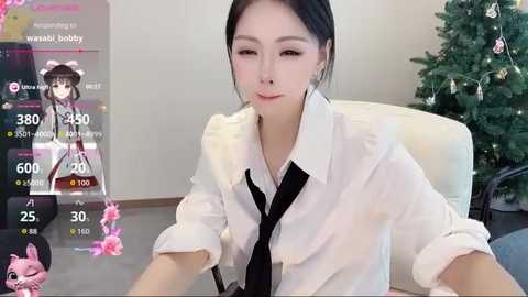 Video of an East Asian woman with straight black hair, wearing a white shirt and black tie, seated indoors near a decorated Christmas tree.