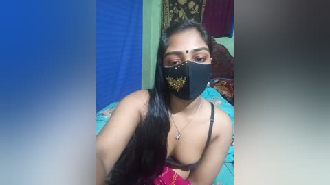 Video of a South Asian woman with long black hair, wearing a black mask, black bra, and turquoise necklace, lying on a bed with floral bedding, in a dimly lit room.