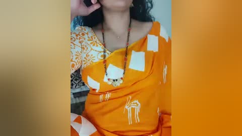 Video of a woman with medium-brown skin, wearing a bright orange saree with white elephant patterns, and a black beaded necklace, holding a phone to her ear.