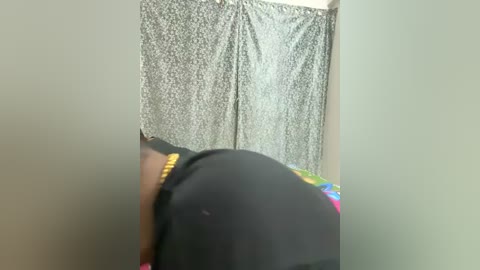 Video of a person wearing a black hat and a colorful shirt, partially visible from the waist down, standing in front of a grey curtain with a floral pattern.