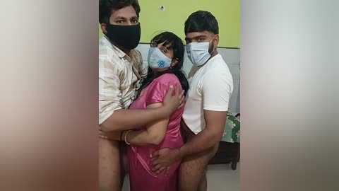 Video of three people: a woman in a pink dress, a man in a striped shirt, and a man in a white t-shirt, all wearing masks. Background shows a yellow wall and a green sofa.