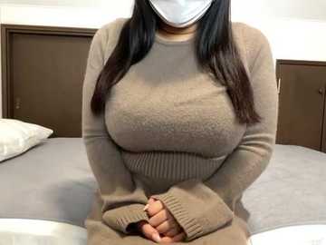 Video of a woman with medium skin tone, wearing a beige long-sleeve sweater, sitting on a bed with white sheets, wearing a white mask, in a simple bedroom setting.