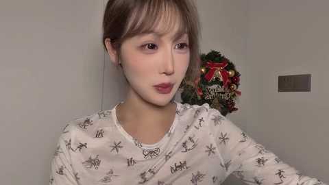 A video of a young Asian woman with fair skin, brown hair, and light makeup. She wears a white shirt with snowflakes. The background features a festive holiday wreath with red and green elements.