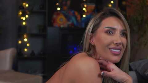 Video of a smiling woman with long blonde hair, medium skin tone, and a slender physique, leaning forward with her elbow on a table, in a dimly lit, colorful room with shelves in the background.