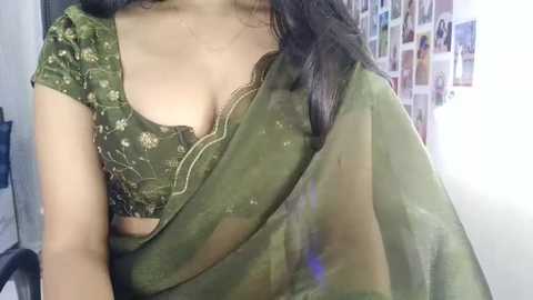 Video of a woman in an olive-green saree with a floral blouse, showcasing her medium-sized breasts. Background features a white wall with a collage of photos.