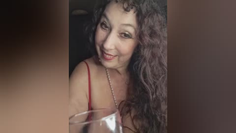Video of a smiling, middle-aged woman with curly dark hair and fair skin, wearing a red spaghetti strap top, holding a wine glass, against a dark background.