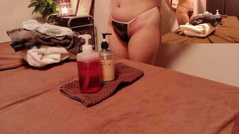Video of a nude woman with pale skin, wearing only a thong, standing in a cluttered bedroom with a mirror.