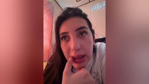 A video of a young woman with long dark hair, light skin, and dark eyebrows, smiling with her finger to her lips. She is indoors with warm lighting and framed artwork in the background.