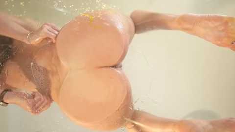 Video of a naked, light-skinned woman with wet, large buttocks and visible pubic hair, standing in a shower, water droplets on her skin, blurry background.