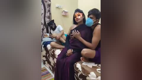 Video of a woman in a purple sari, wearing a blue mask, sitting on a bed with a patterned bedspread. A man in a white shirt and black mask sits nearby, another woman in a black sari is also present.