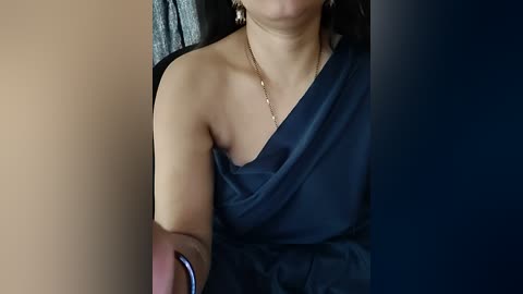 Video of a woman with light brown skin, wearing a navy blue, off-the-shoulder dress and a delicate gold necklace, sitting on a dark chair, background blurry.