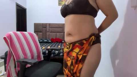 Video of a light-skinned, plus-sized woman with medium-sized breasts, wearing a black bra and colorful patterned pants, standing in a simple, modern bedroom with a striped chair and a bed with checkered bedding.