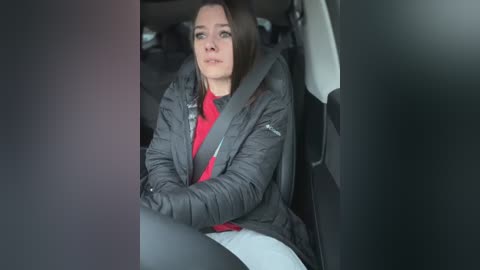 Media: Video of a woman with shoulder-length brown hair, wearing a gray puffer jacket and red shirt, sitting in a car seat, looking serious, with a black car door partially visible.