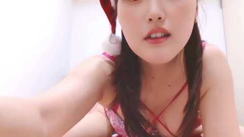 Video of an Asian woman with fair skin, long dark hair, and a Santa hat. She wears a red lace bra, with a subtle makeup look. The background is blurred, focusing on her.