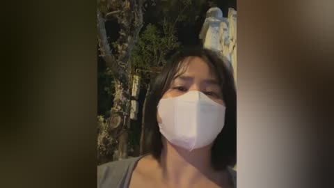 Media: Video of an Asian woman with straight, shoulder-length black hair, wearing a white surgical mask and a gray shirt, against a dimly lit, cluttered background with green plants and white objects.