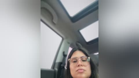 Video of a woman with long dark hair, wearing glasses, sitting in a car with a sunroof, looking down at a phone.