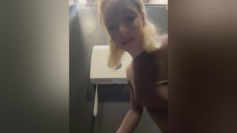 A blurry video of a blonde woman in a public restroom, wearing a light-colored top, leaning over a white toilet. The background features a gray wall and a dispenser.