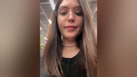 Video of a young woman with long, straight, light brown hair, wearing round glasses and a black top. She has a light complexion and a neutral expression. The background is blurred with beige walls and a ceiling.