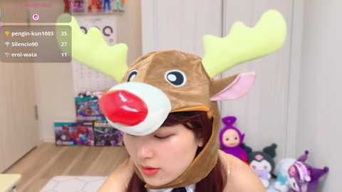 A young woman with fair skin, wearing a brown reindeer costume with large antlers, red nose, and brown fur, poses indoors with a wooden floor. The background shows a shelf with toys and a stuffed purple creature.