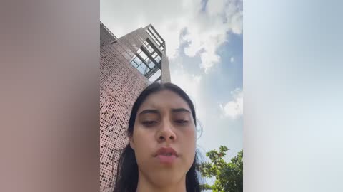 Video of a young woman with long, straight black hair, standing against a brick wall with a modern glass building and a partly cloudy sky in the background.
