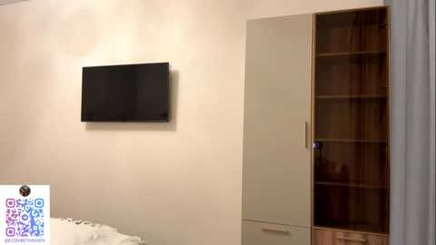 Video of a minimalist bedroom with a beige wall, a flat-screen TV mounted above a bed with white sheets, and a tall, beige wardrobe with wooden shelves.