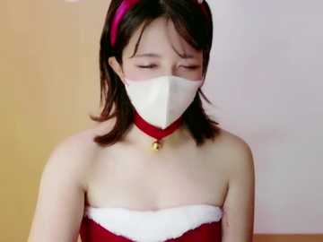 Video of an Asian woman with shoulder-length dark hair, wearing a red Santa hat, white face mask, and a red strapless dress with a white collar, against a gradient background.