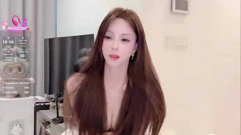 Video of a fair-skinned Asian woman with long brown hair, wearing a low-cut black dress, standing in a modern, brightly lit room with white walls and a large TV screen.