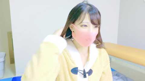 Video of a young Asian woman with fair skin, wearing a yellow hoodie, sitting in a room with a white wall and a wooden bed. She is holding a pink mask to her mouth, looking slightly upset.