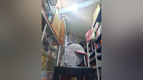 Video of a narrow, dimly-lit clothing store with glass display cases, filled with colorful, neatly organized garments, including a woman wearing a white, patterned headscarf, standing on a small stool.