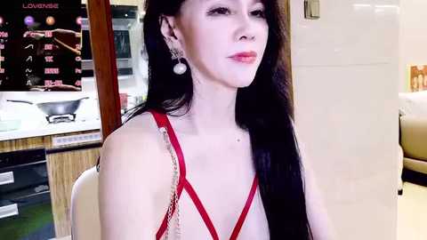 Video of a slender, fair-skinned Asian woman with long black hair, wearing a red halter dress, seated indoors. Background features a kitchen and a partially visible dining area.