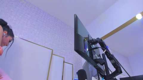 Video of a woman with black hair wearing a pink top, standing in a room with a white brick wall and a large, black monitor on a stand, reflecting a blue light.