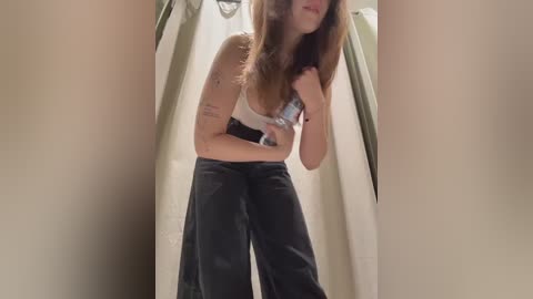Video of a young woman with long brown hair, wearing a light blue bra and baggy black pants, standing in a shower with beige curtains.